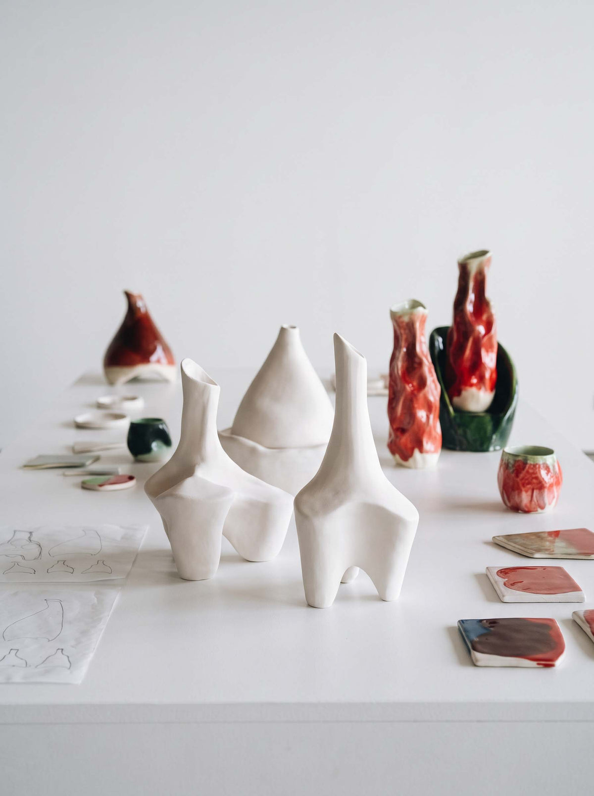 Calassi ceramic wine vessel prototypes in white, red and green, displayed with sketches and glaze samples on a white studio table