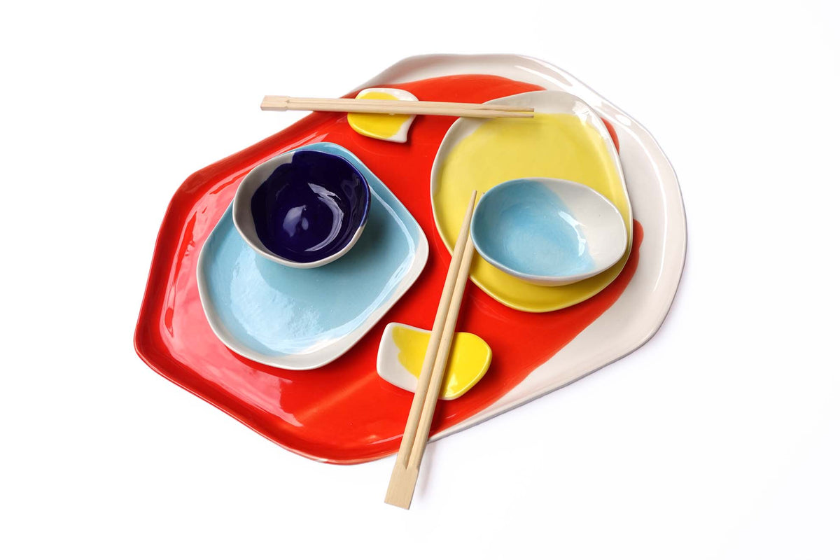 Stacked sushi set with a red tray, yellow and light blue plates, dip bowls and wooden chopsticks