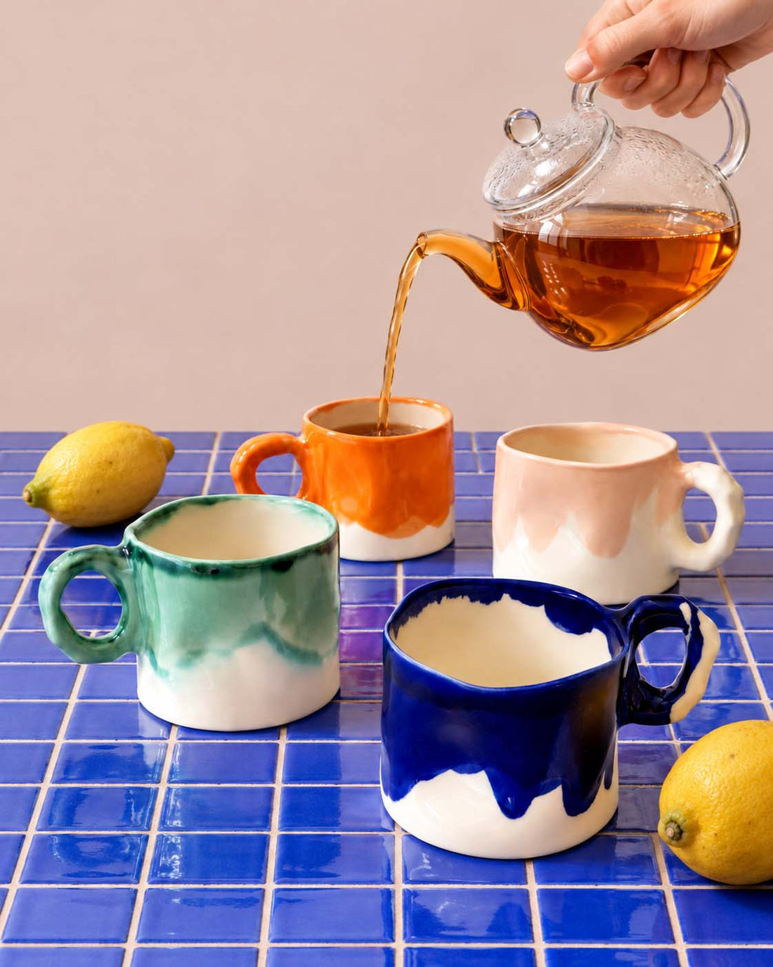 Tea poured into colourful handmade mugs beside lemons on a blue tiled table