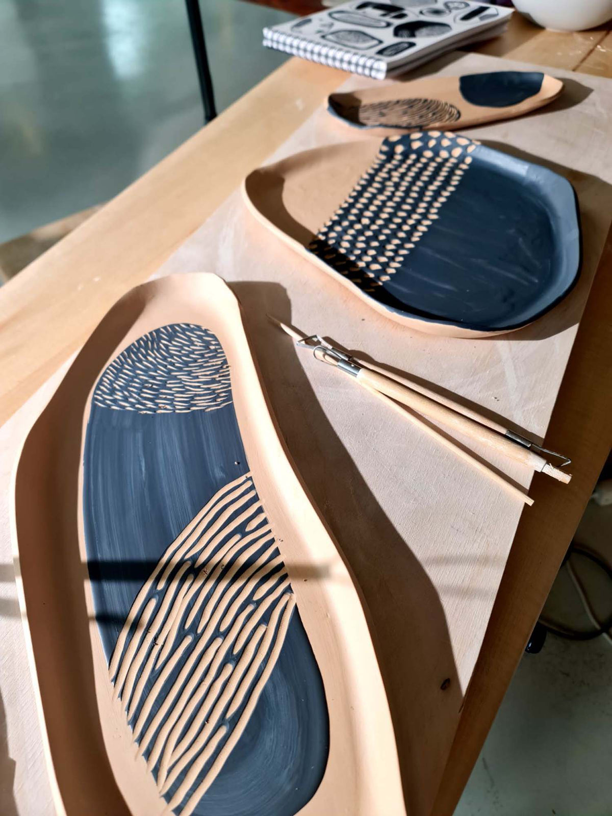 Organic-shaped sgraffito platters with blue glaze and carved line patterns on a studio table