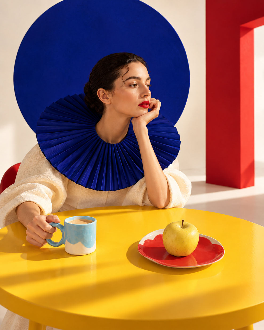 Woman with a sculptural blue collar beside a sky blue ceramic mug and a yellow apple on a red plate