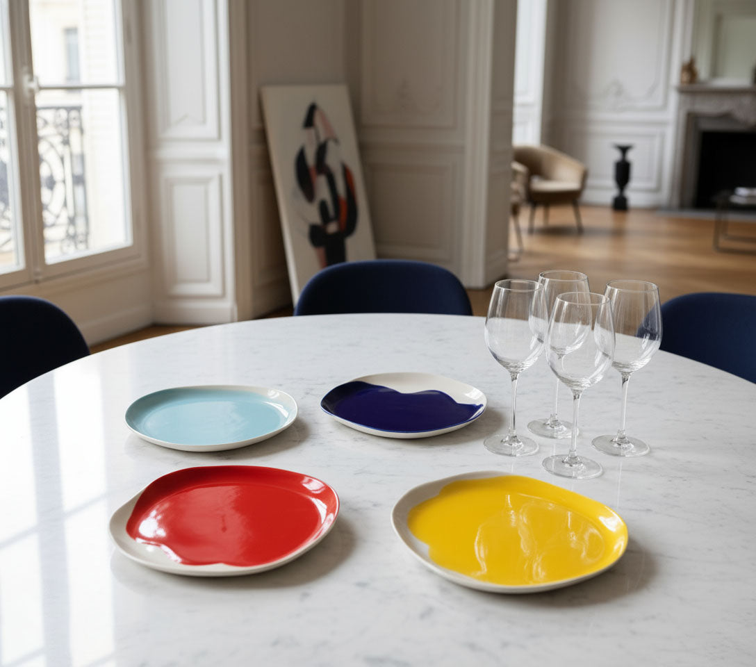 Four colourful BAUHAUS ceramic plates and wine glasses on a pale dining table in an interior