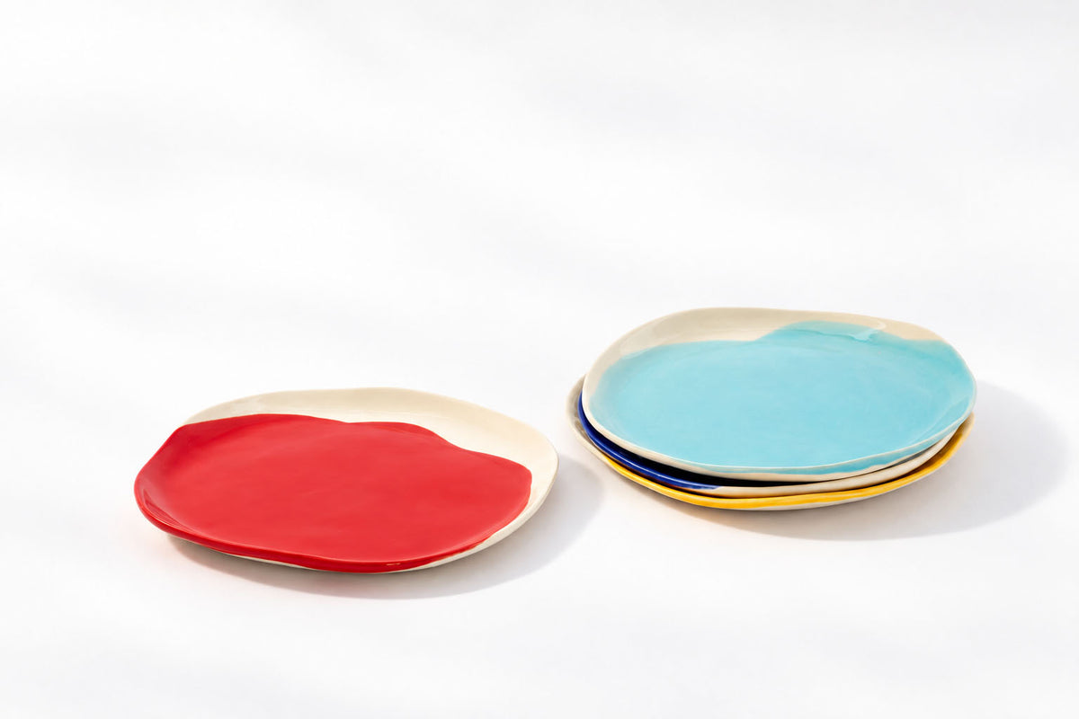 Red BAUHAUS ceramic plate beside stacked sky blue, cobalt blue and yellow plates on white