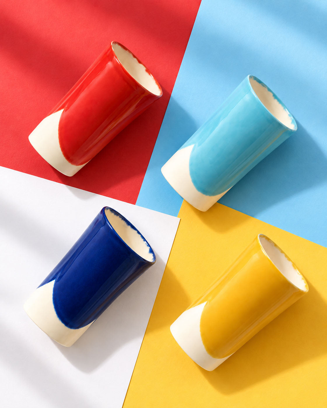 Four BAUHAUS ceramic tumblers in red, sky blue, cobalt blue and yellow lying on coloured paper