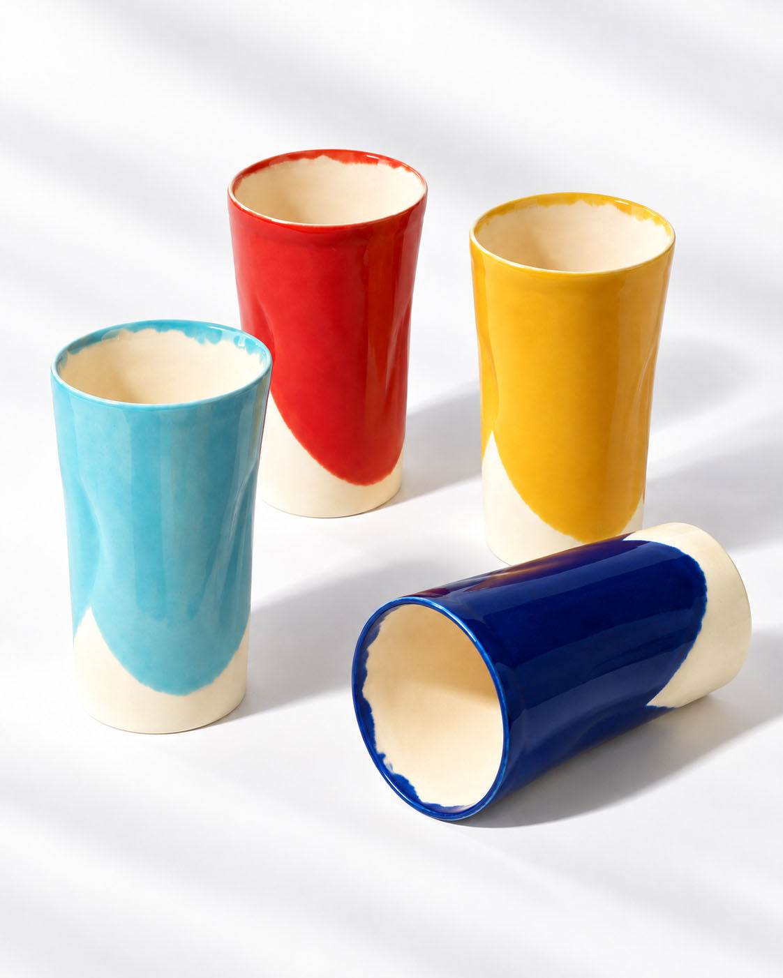 Four BAUHAUS ceramic tumblers on white, with the cobalt blue tumbler lying beside the red, yellow and sky blue cups