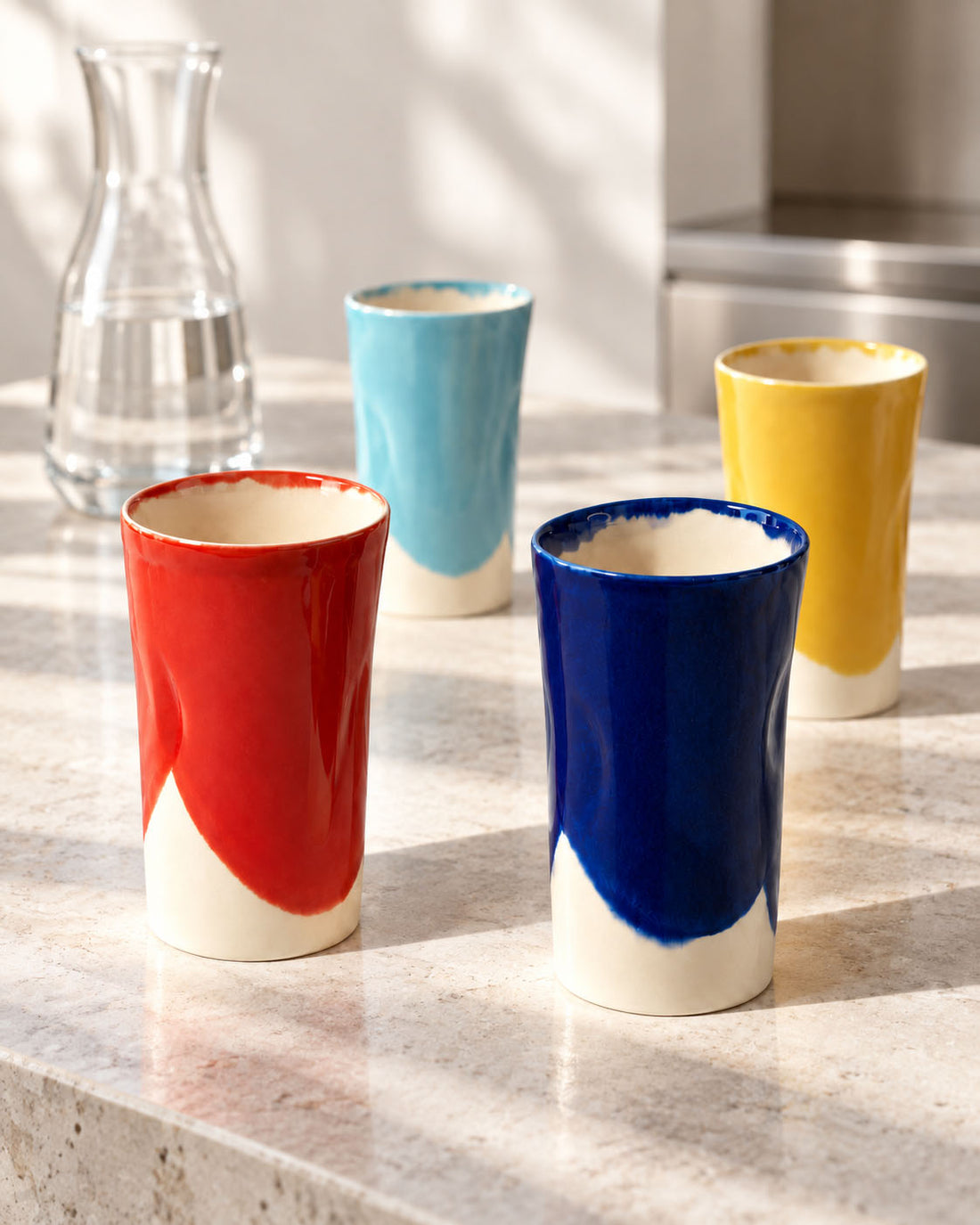 Four colourful BAUHAUS ceramic tumblers on a pale stone counter beside a glass water carafe