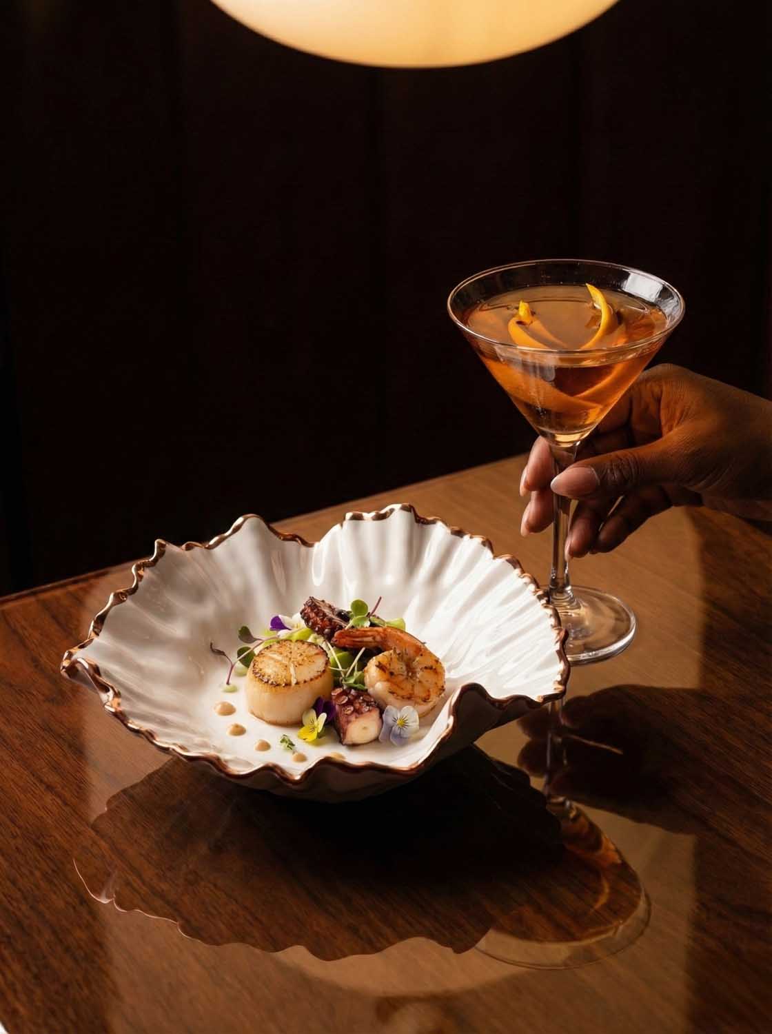 Scallop dish on a ruffled white porcelain plate beside a cocktail glass in a dim restaurant
