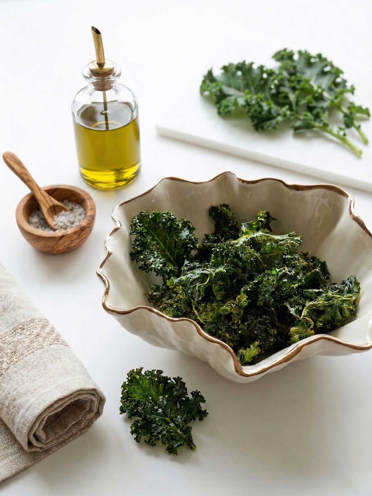 Blossom bowl with copper rim filled with kale chips, beside olive oil and a wooden salt bowl