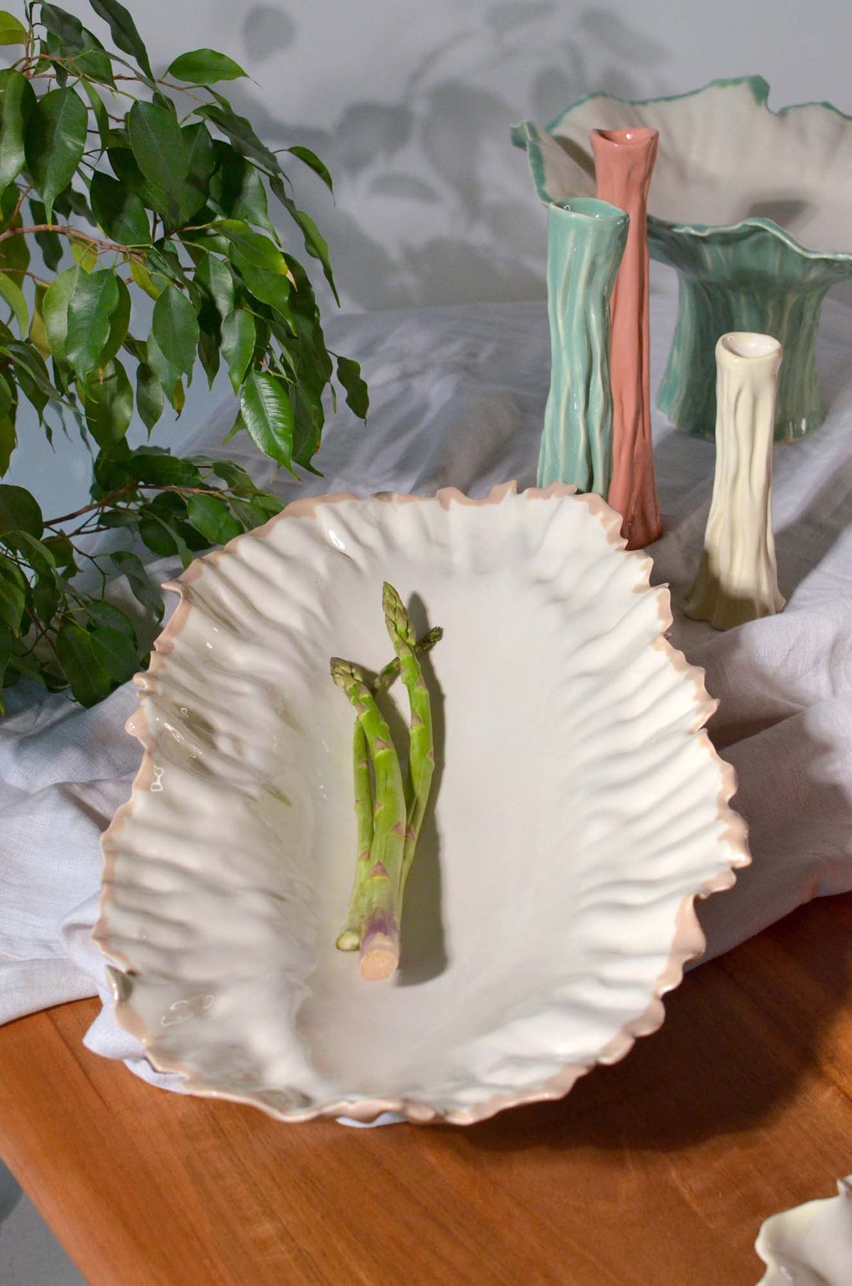Blossom elevated serving bowl with green asparagus on a wooden table beside Blossom vases