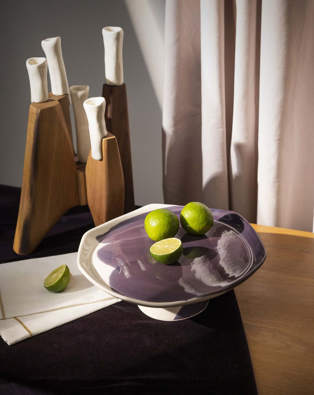 Porcelain cake stand in Aubergine glaze holding limes, set on a dark dining table