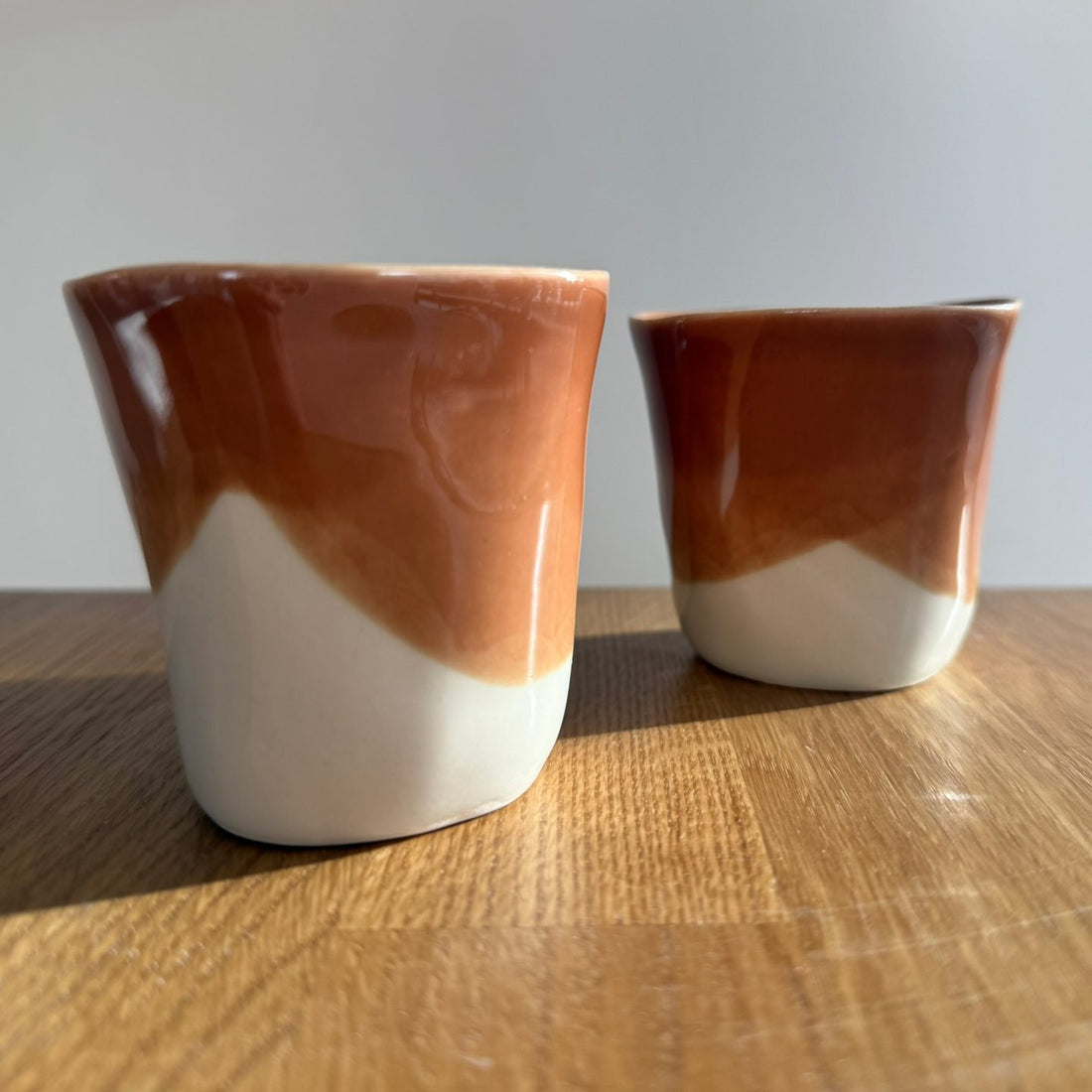 Two handmade porcelain cups dipped in Hazelnut glaze over white on a wooden table