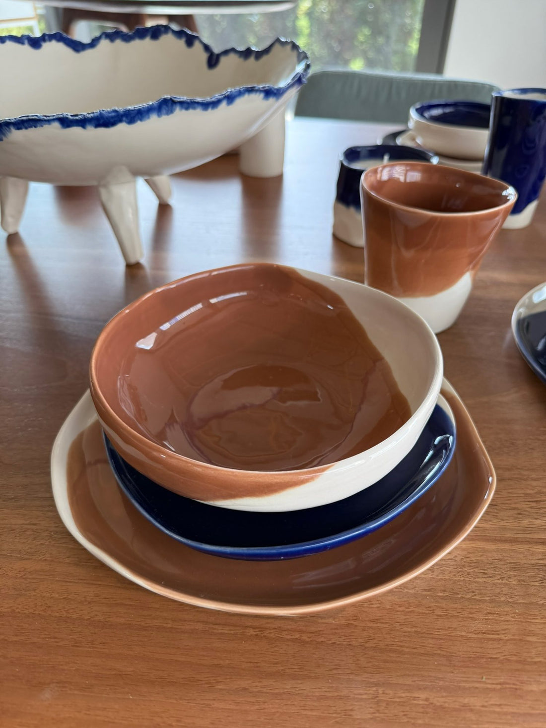 Hazelnut glazed porcelain bowl on a navy plate, with cups and a footed bowl on a wooden table