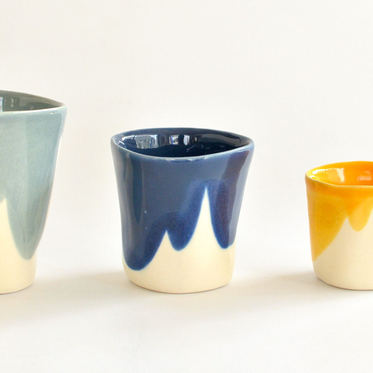 Handmade porcelain cup with dripping Dark Denim glaze beside light blue and yellow cups