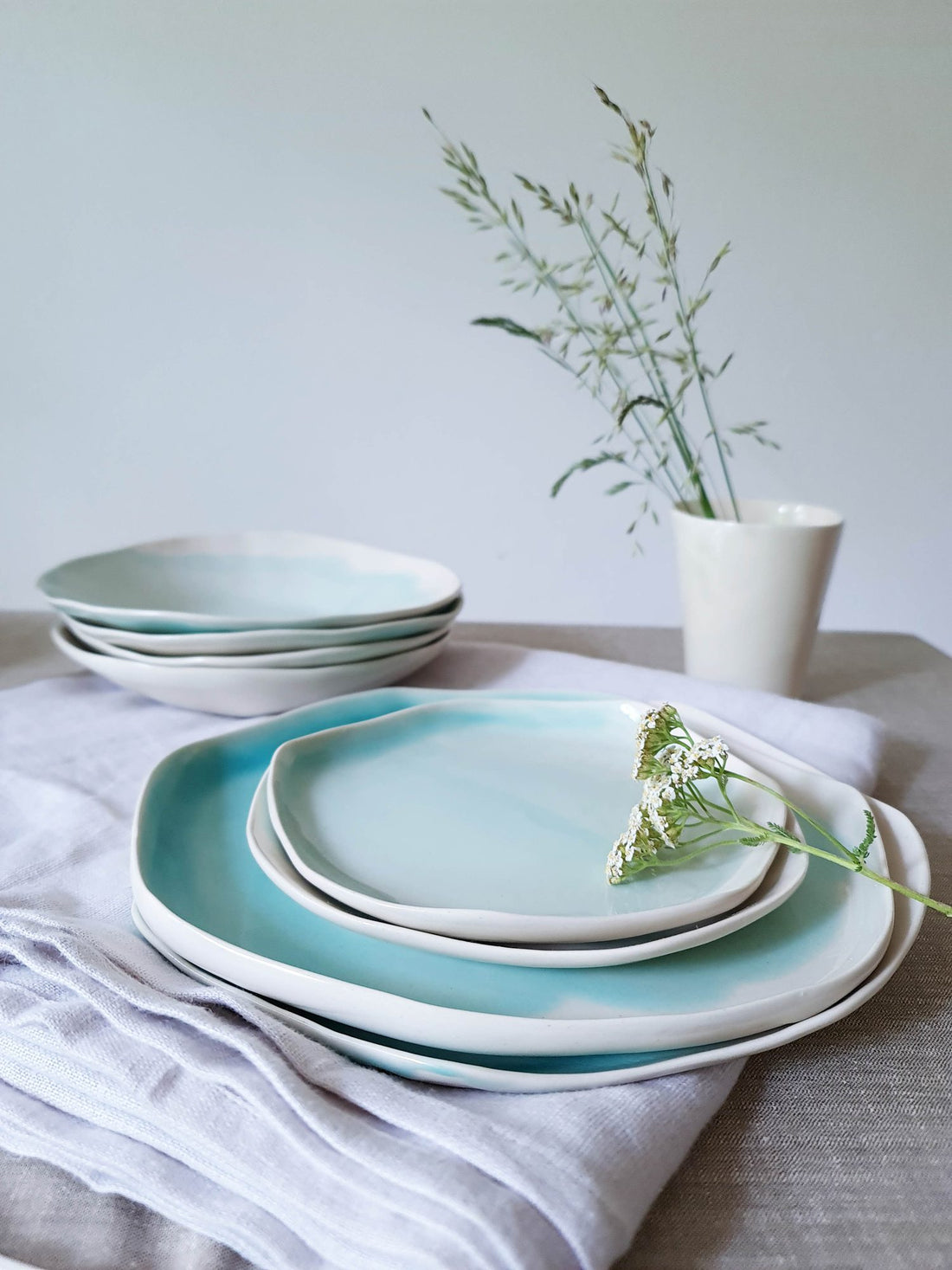 Turquoise glazed handmade ceramic dinnerware stacked on linen with wild grasses in a vase