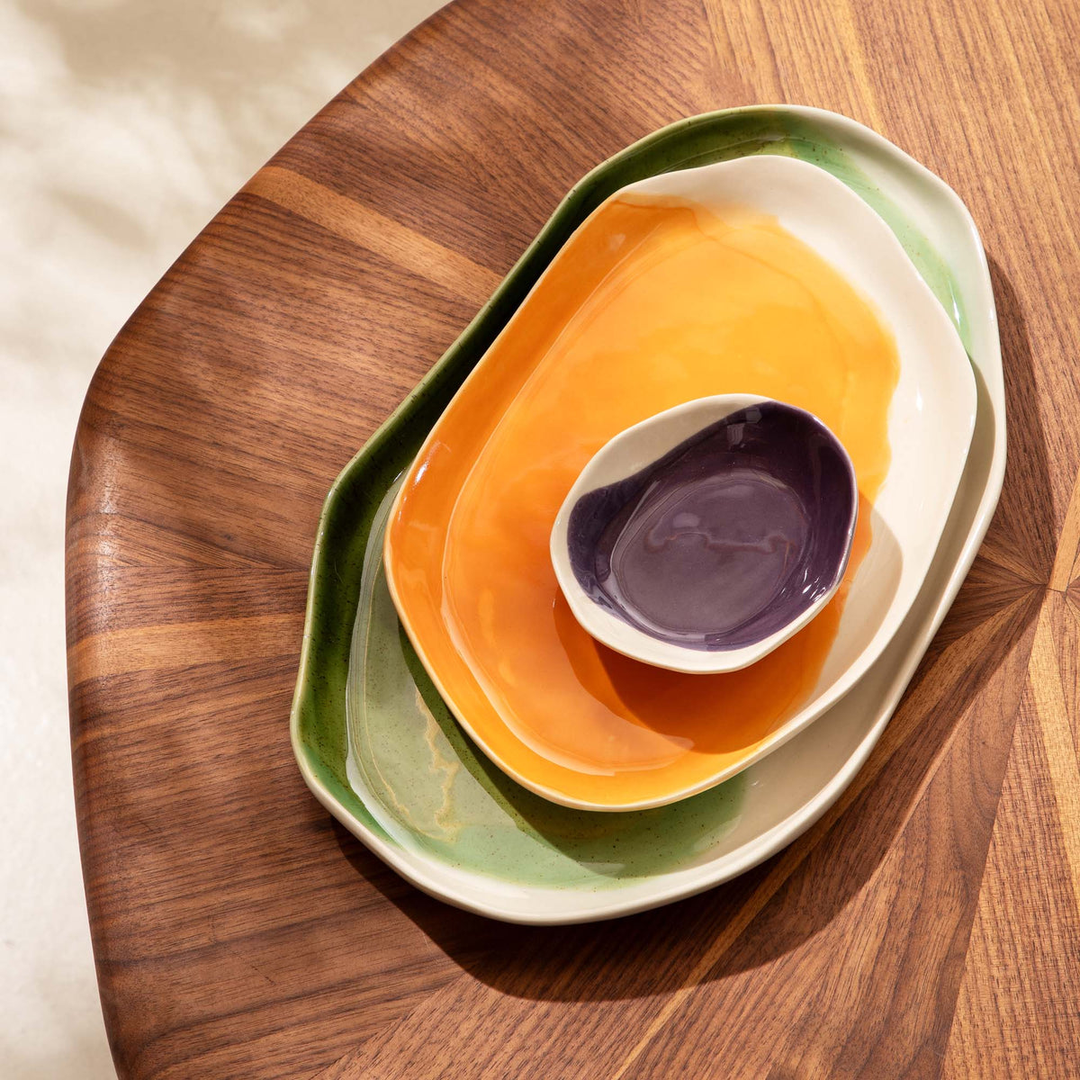 Nested handmade serving trays in green and orange glaze with a purple dip bowl on a wooden board