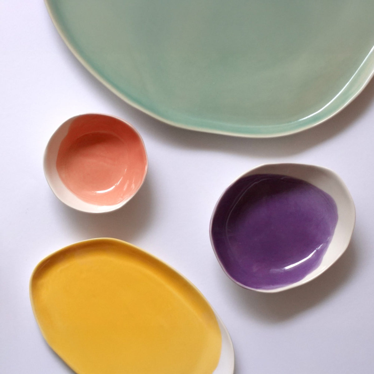 Handmade dip bowls in coral and purple glaze in two sizes, next to a yellow tray and a mint platter