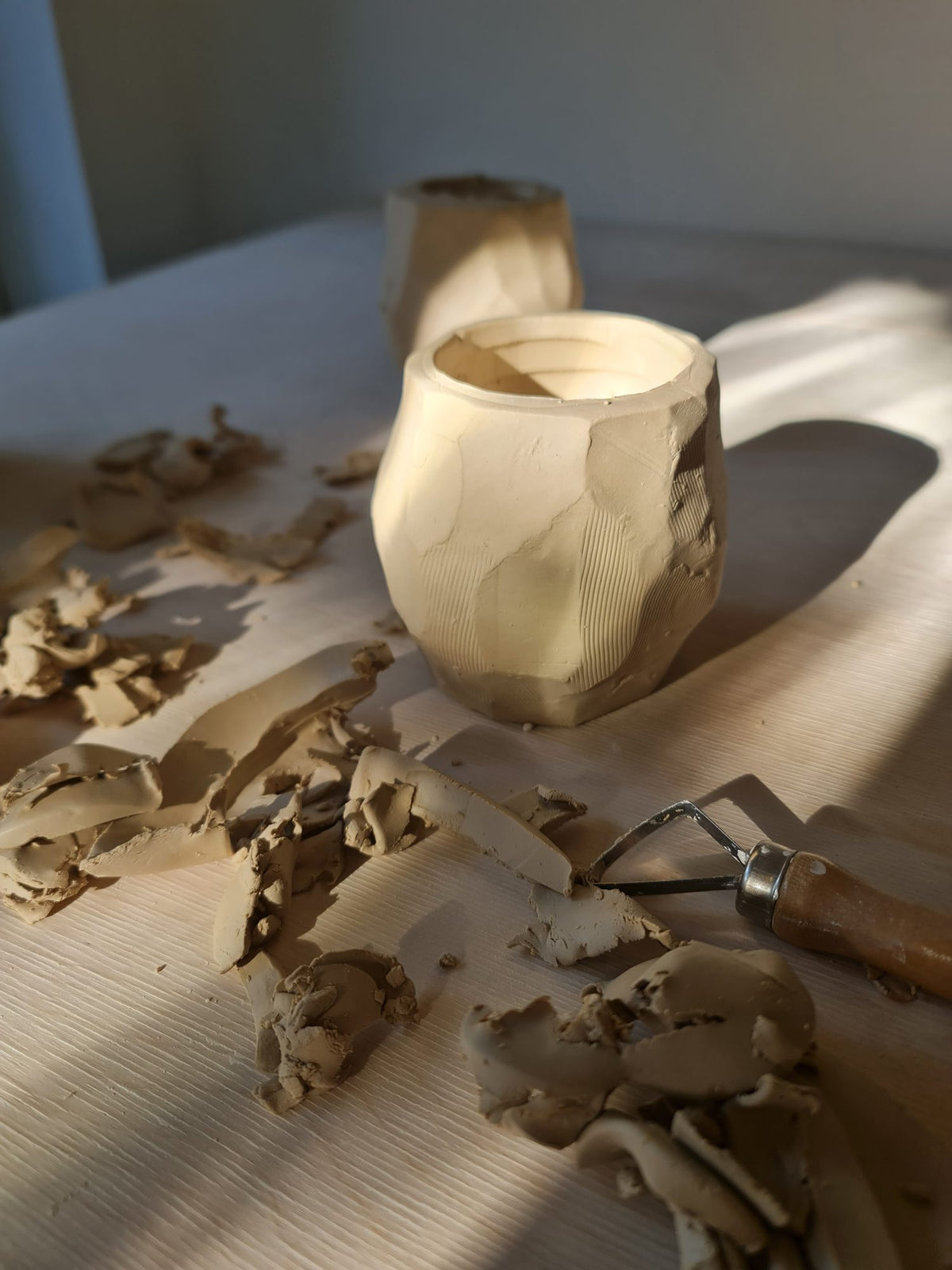 Hand-carved kurinuki clay cup surrounded by clay shavings and a carving tool on a wooden table