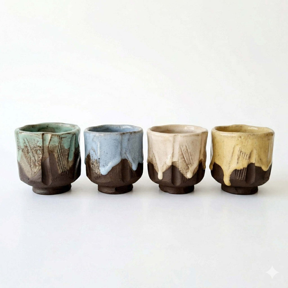 Row of four handmade kurinuki coffee cups with green, blue, cream and yellow glaze on dark clay