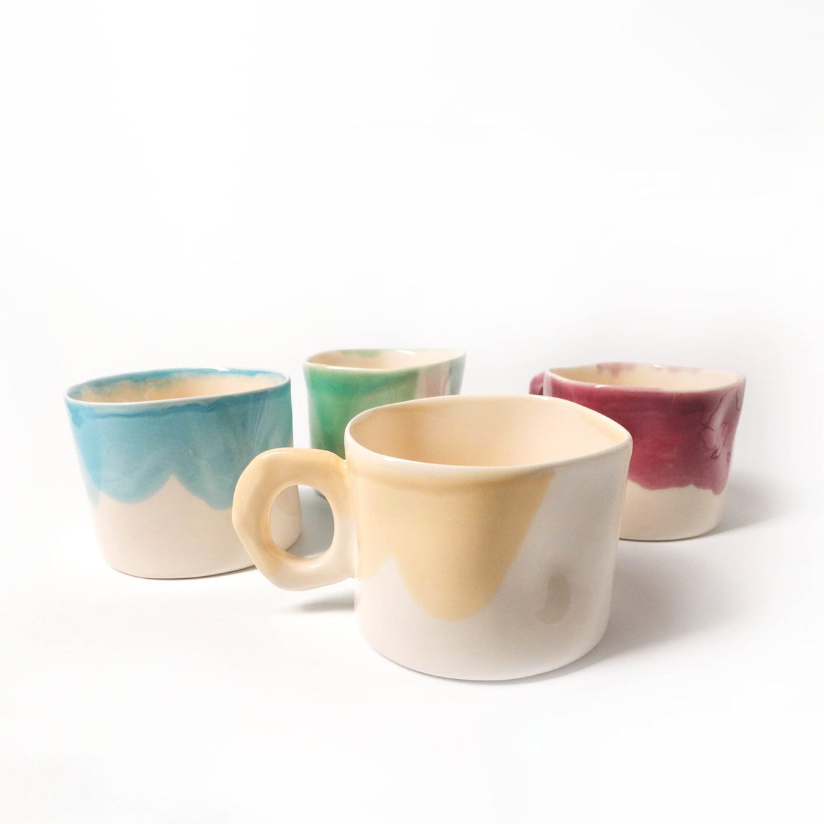 Four handmade mugs with blue, green, pink and yellow glazes on a white background
