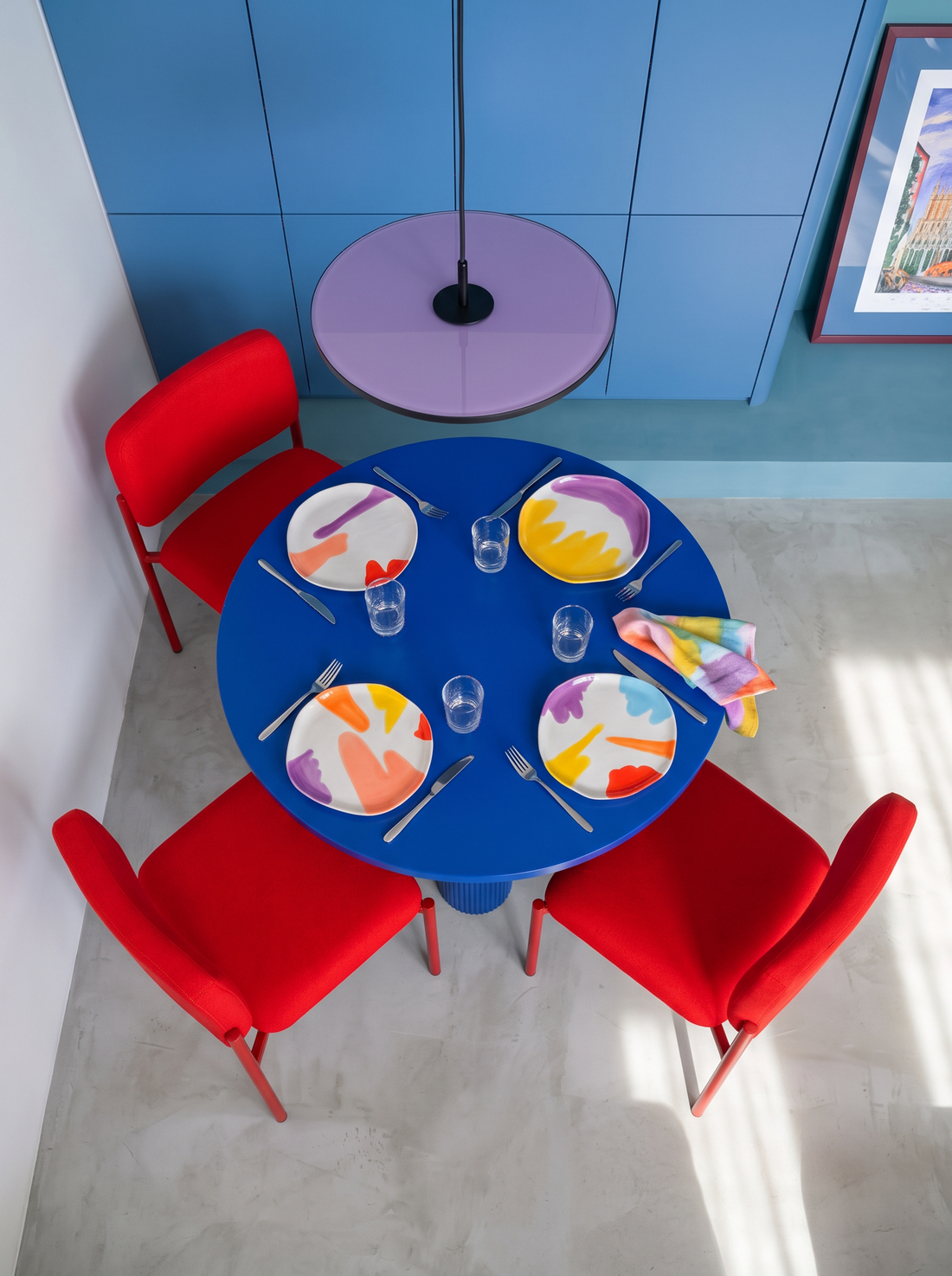 Four colourful ceramic plates arranged on a round blue dining table with red chairs in a contemporary interior