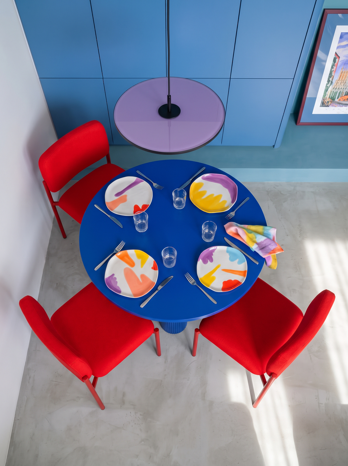 Four colourful ceramic plates arranged on a round blue dining table with red chairs in a contemporary interior