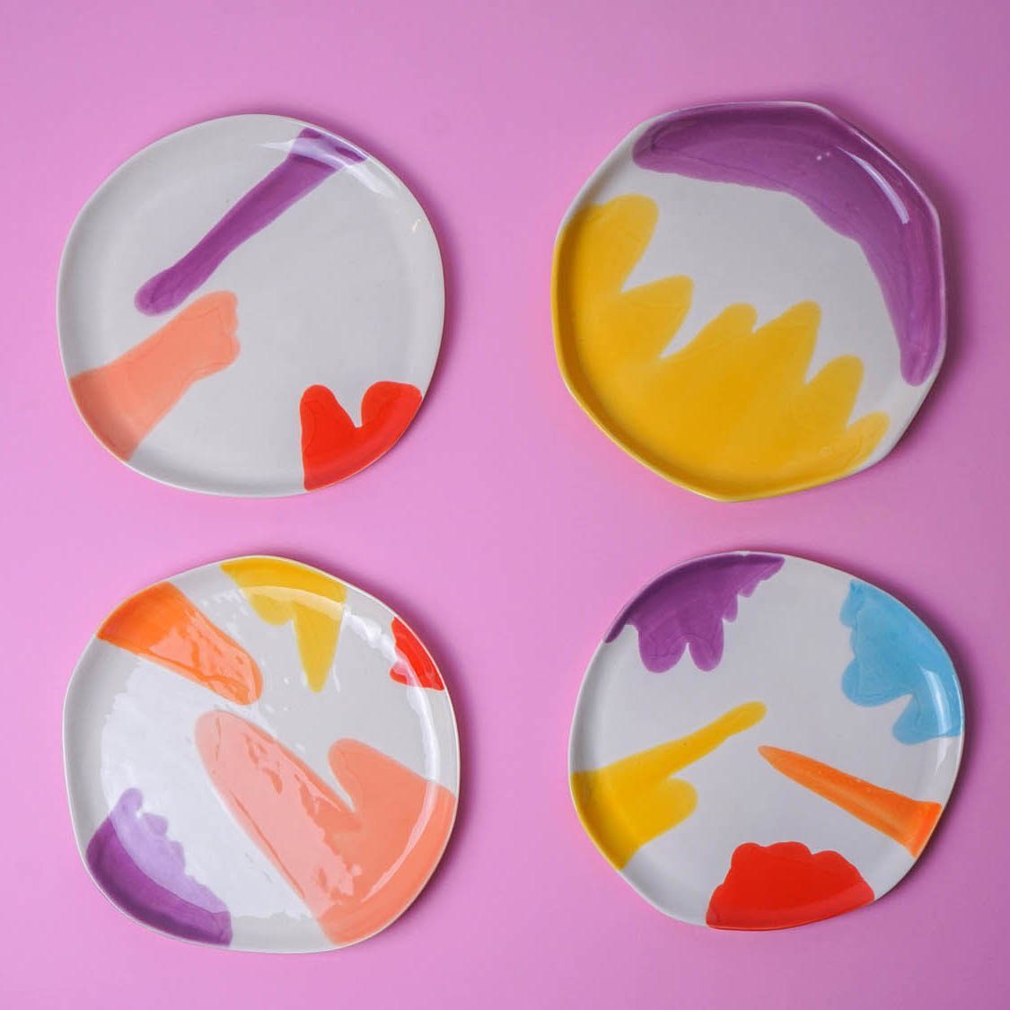 Four ceramic plates with colourful brushstroke designs in purple, yellow, coral, red and blue on a pink background