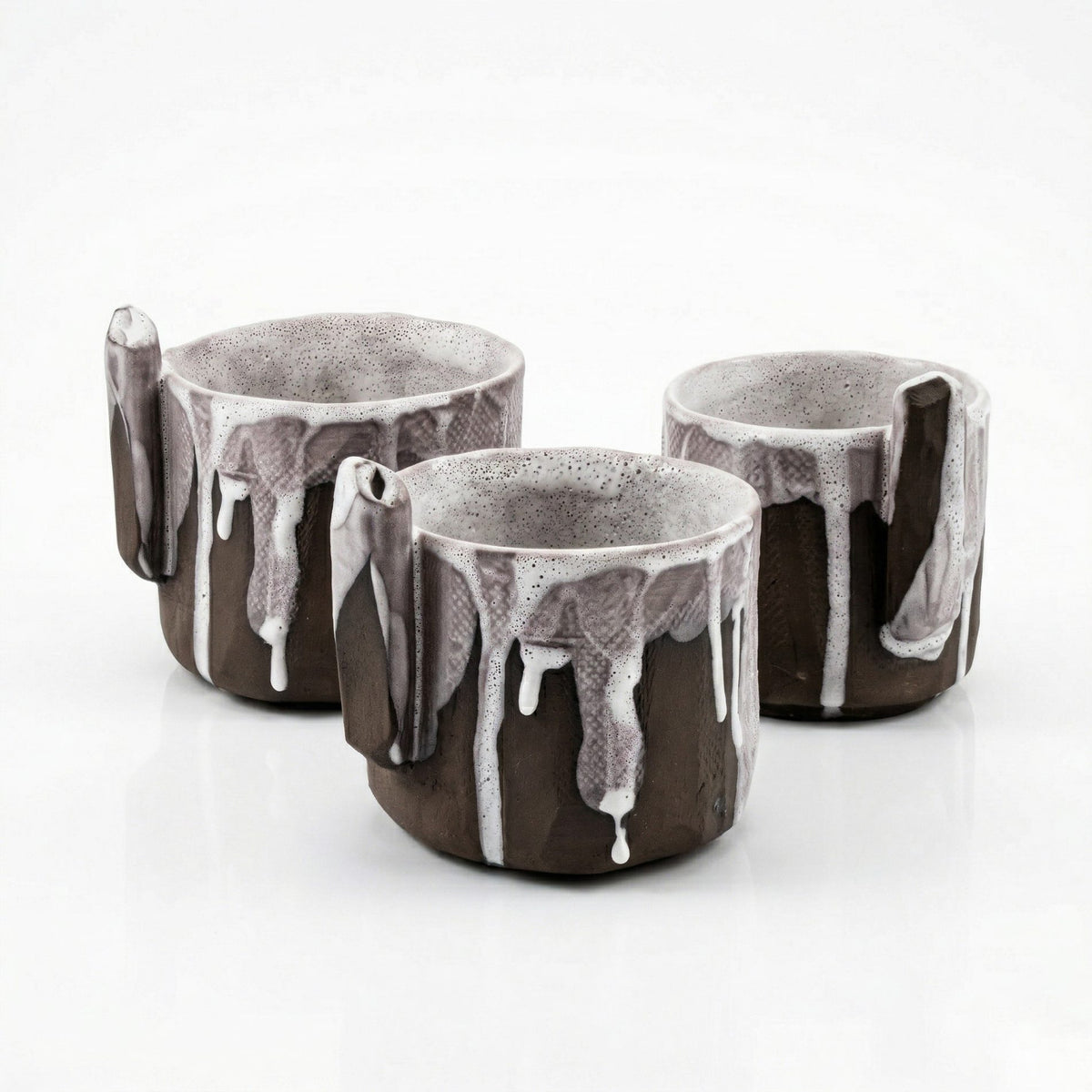 Three handled black clay cups with dripping white glaze on a white background