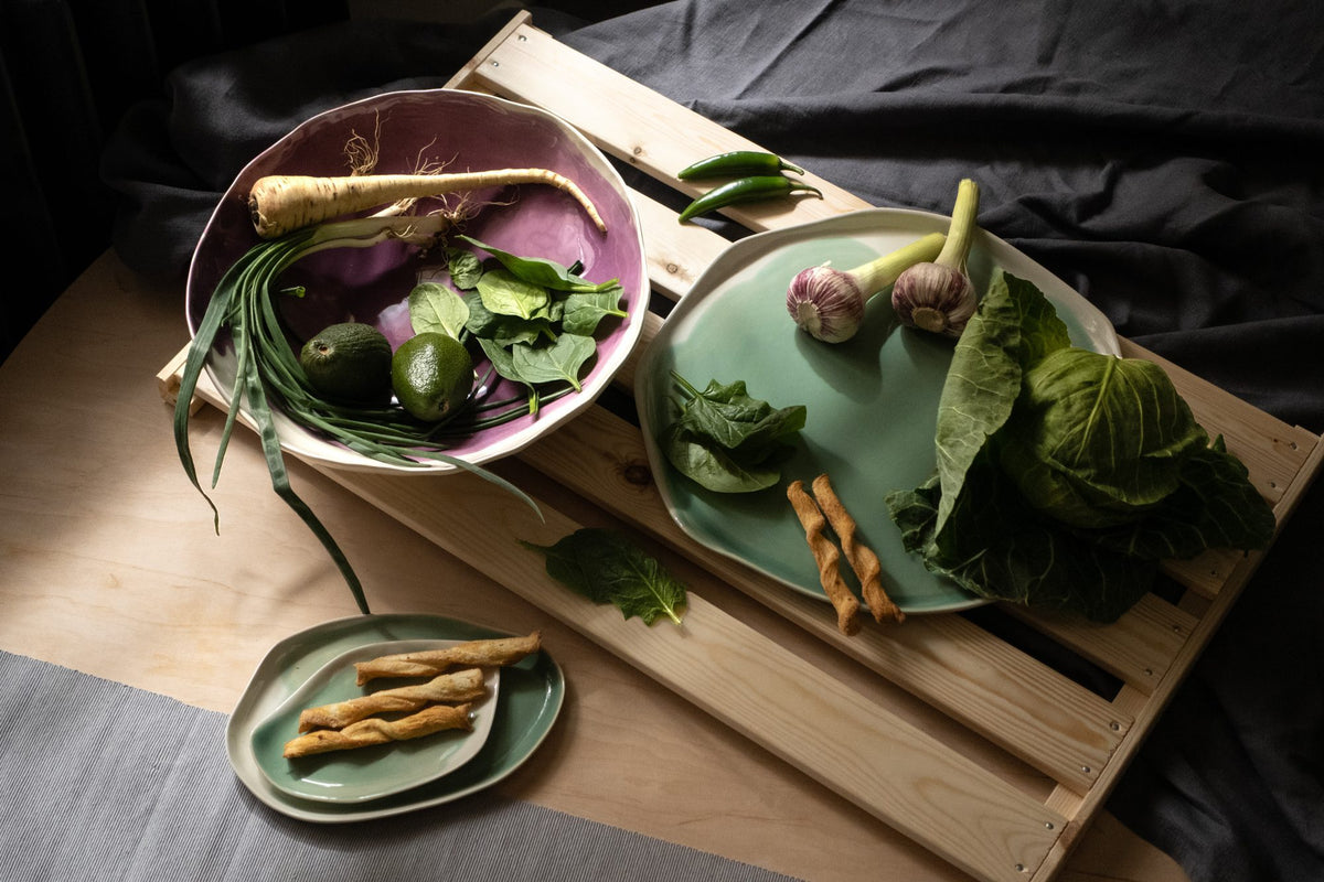 Purple porcelain serving bowl with vegetables next to sage green plates on a wooden crate