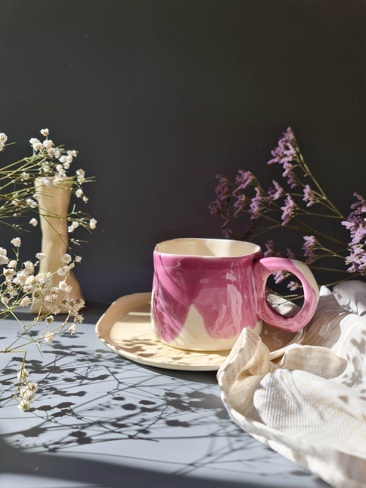 Pink glazed handmade mug on a patterned tablecloth beside dried flowers