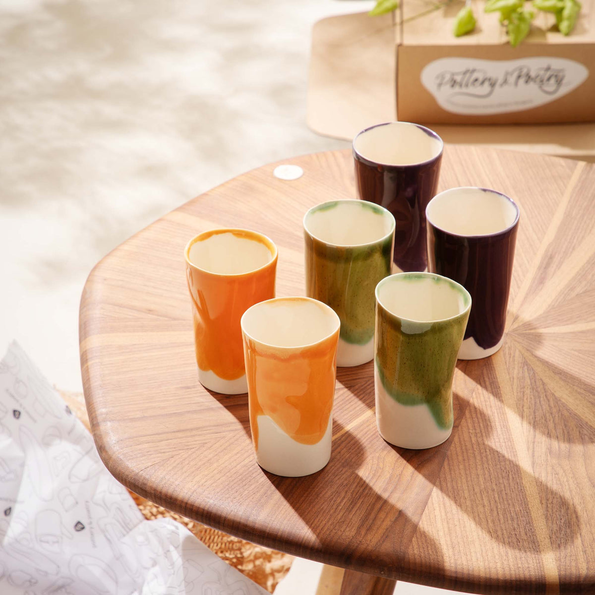 Six tall handmade ceramic cups in orange, green and aubergine glazes on a wooden table