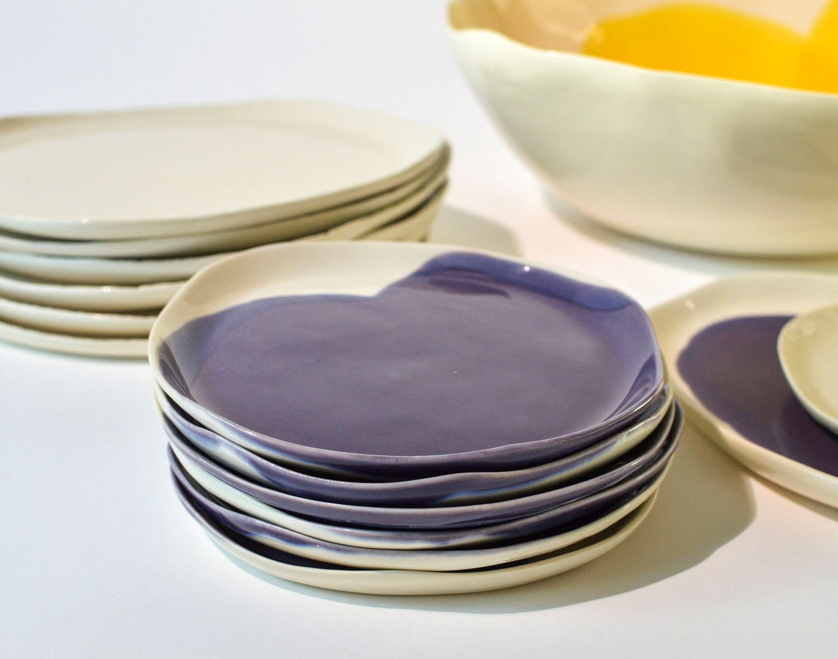 Stack of handmade porcelain side plates in Aubergine glaze beside ivory plates and serving bowls