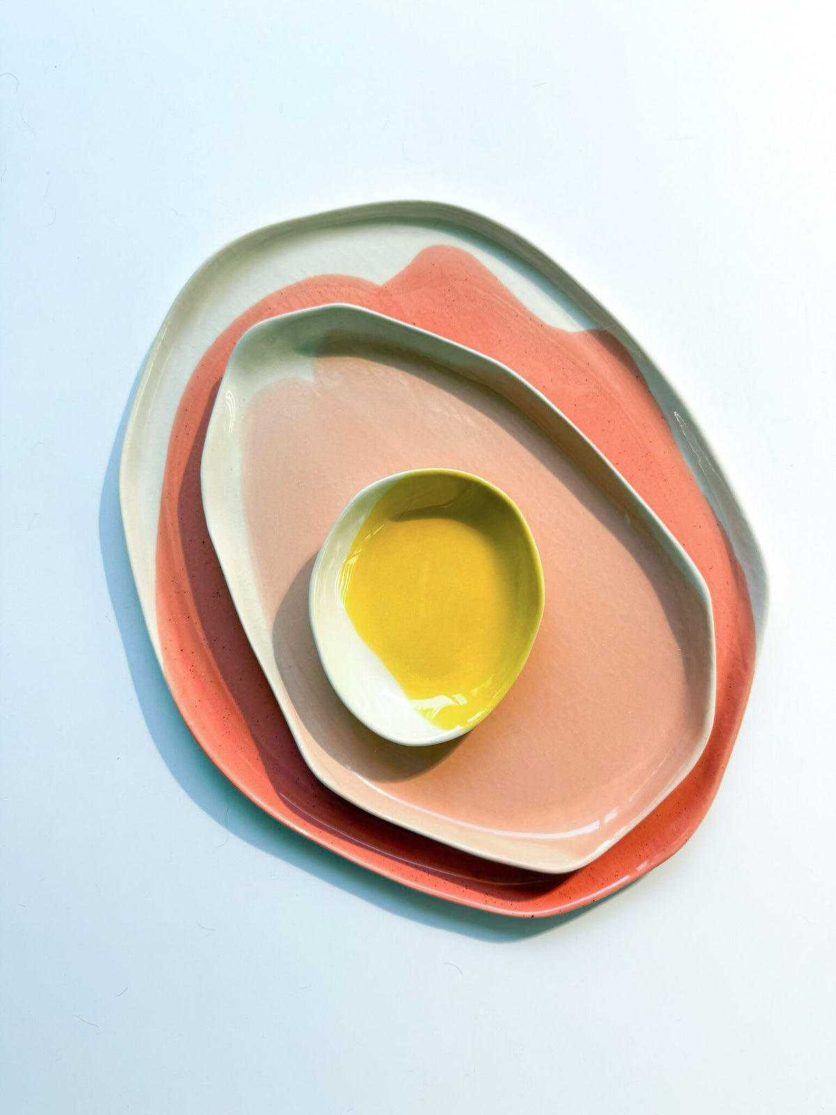 Set of three nested porcelain trays in coral, pink and yellow glaze on a light blue background