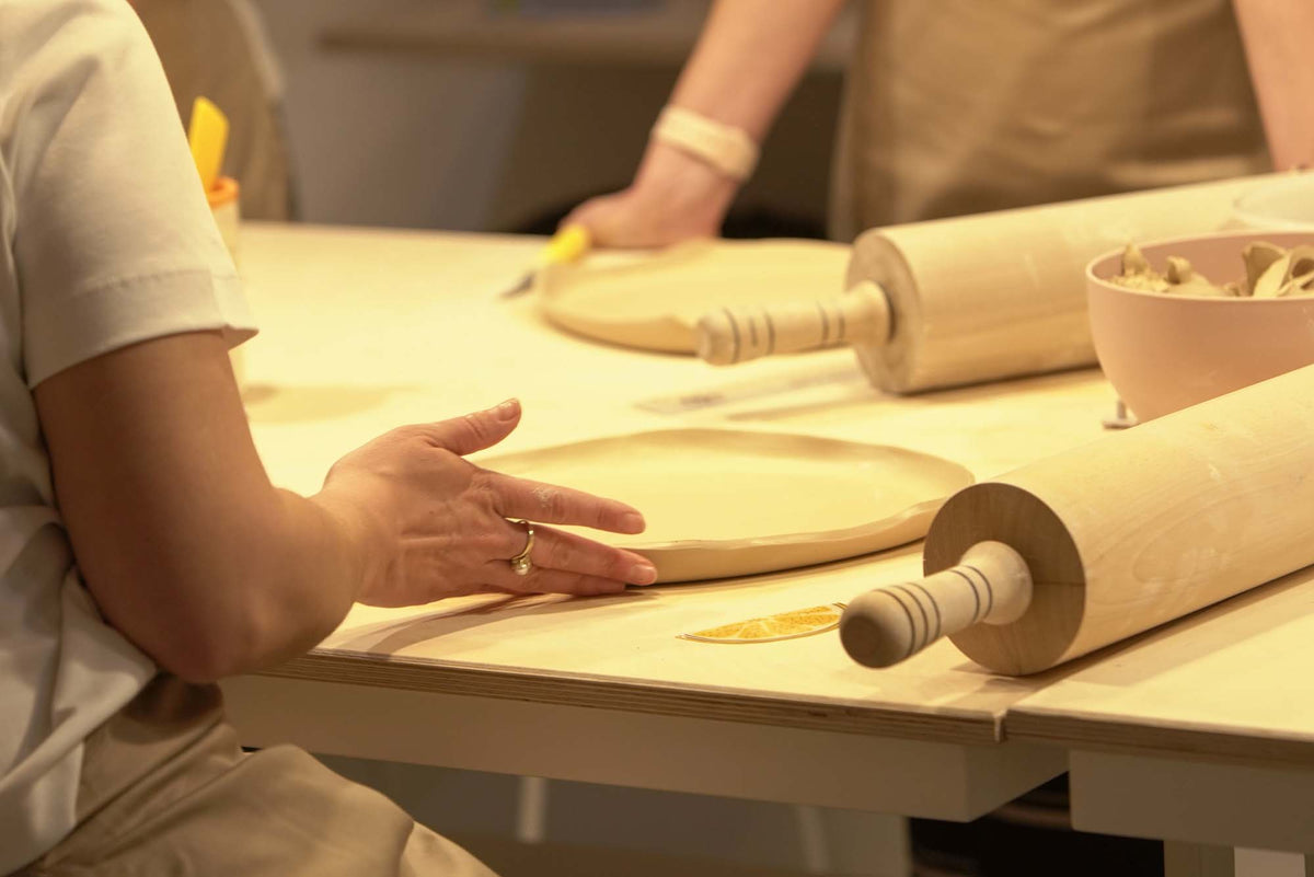 Hands pressing a clay slab into a plate shape next to rolling pins during a pottery workshop