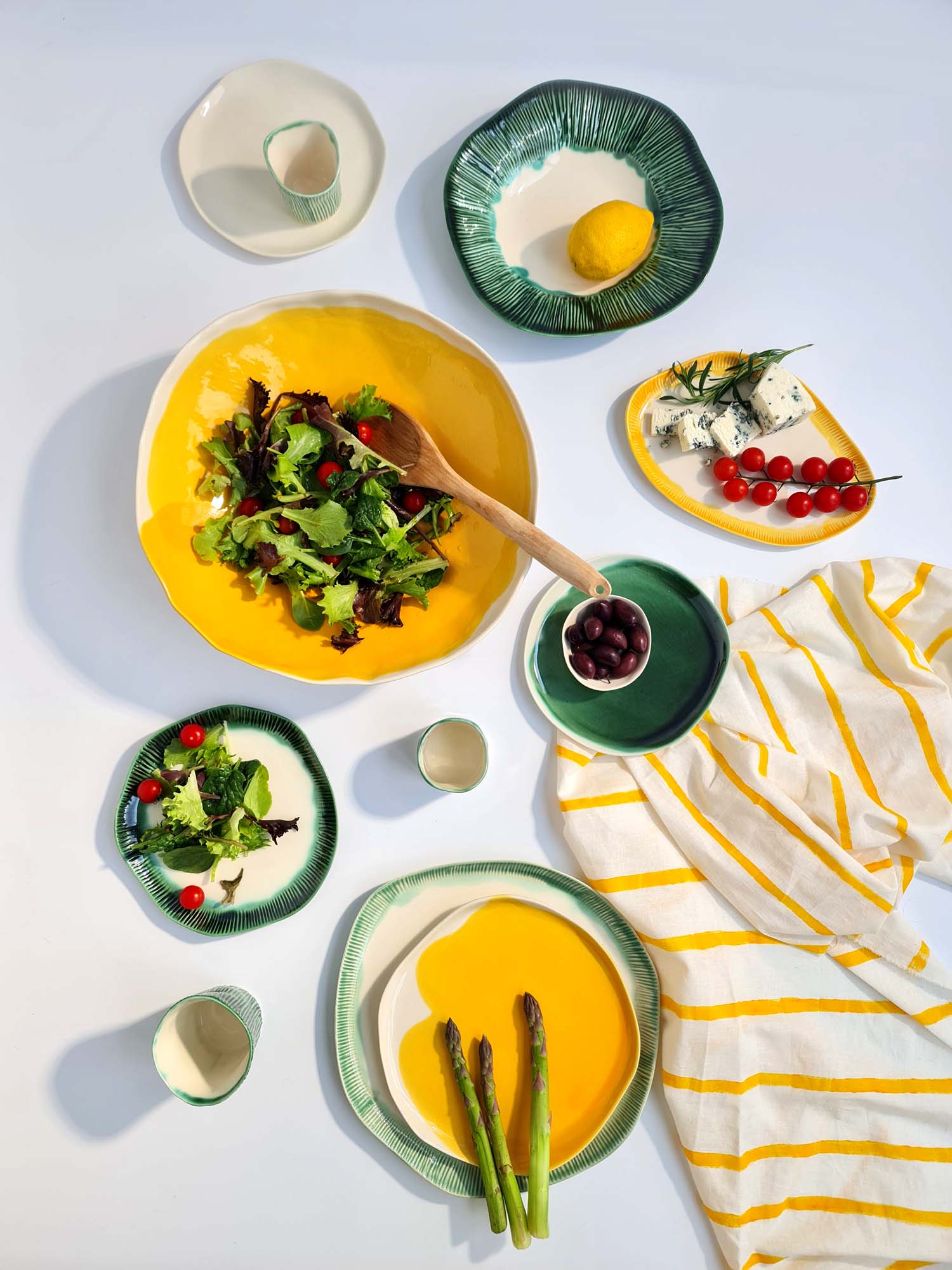 curate your table story (2) Colorful porcelain tableware with salad, asparagus, cheese, and lemons, styled on a white background with a yellow-striped cloth.