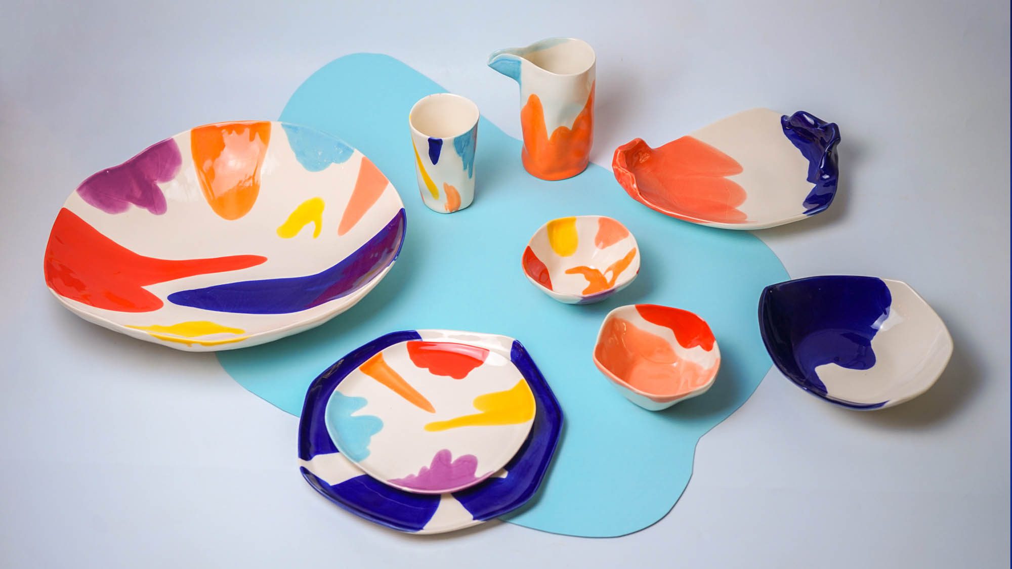 multicolord collection (2) Colorful porcelain plates, bowls, and cups with multicolored brushstroke designs on a light blue background.