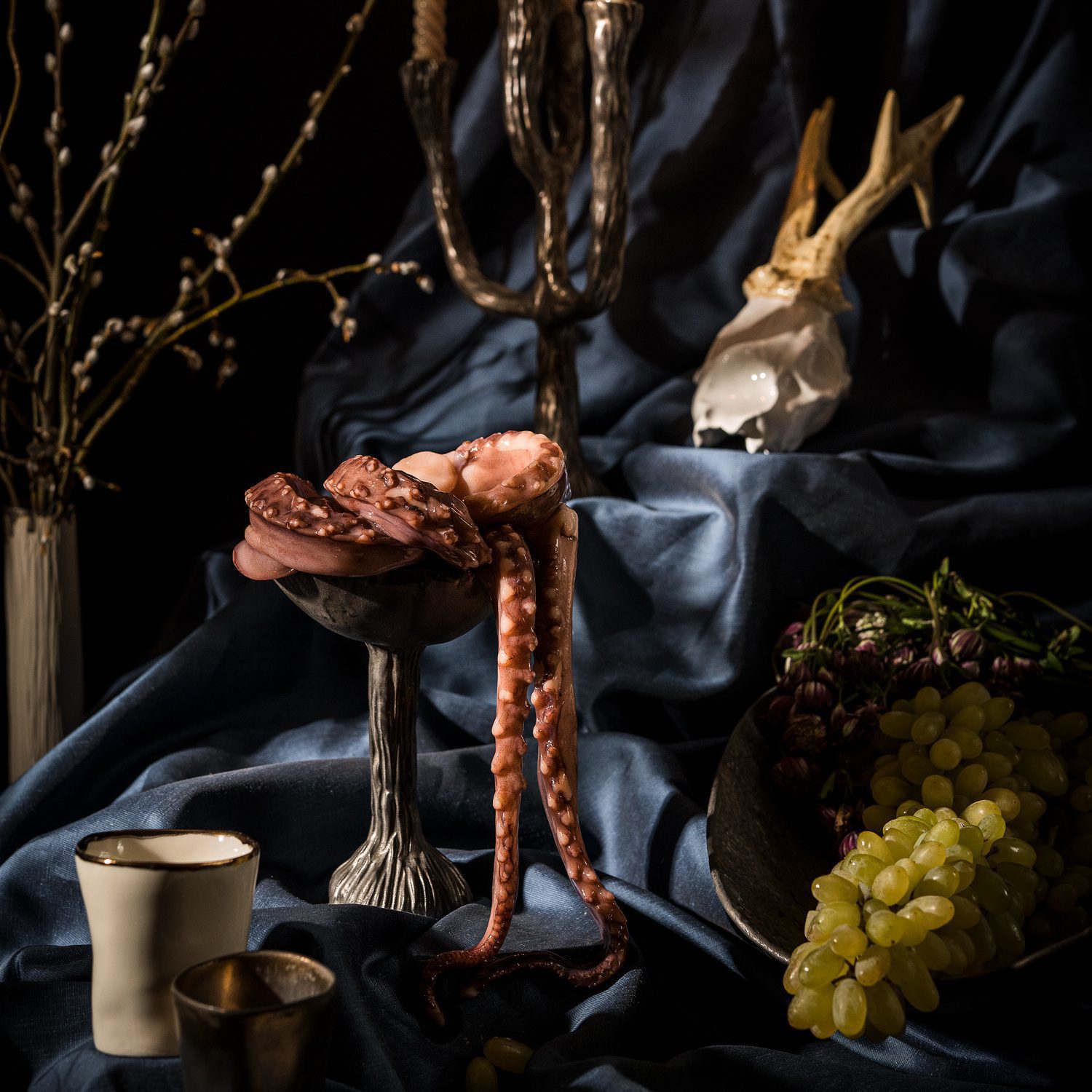 terra lumina set 1 Dramatic still life with octopus tentacles on a sculptural ceramic pedestal, grapes, candles, and dark drapery.